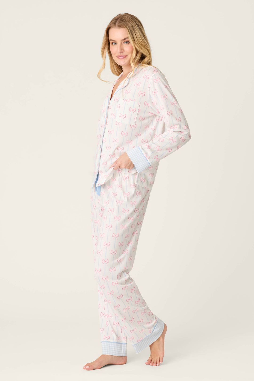 A person stands barefoot in a P.J. Salvage Playful Prints All Wrapped Up Long PJ Set, featuring bow prints and light blue checkered cuffs. The button-front shirt and matching pants create a charming look against the plain background.