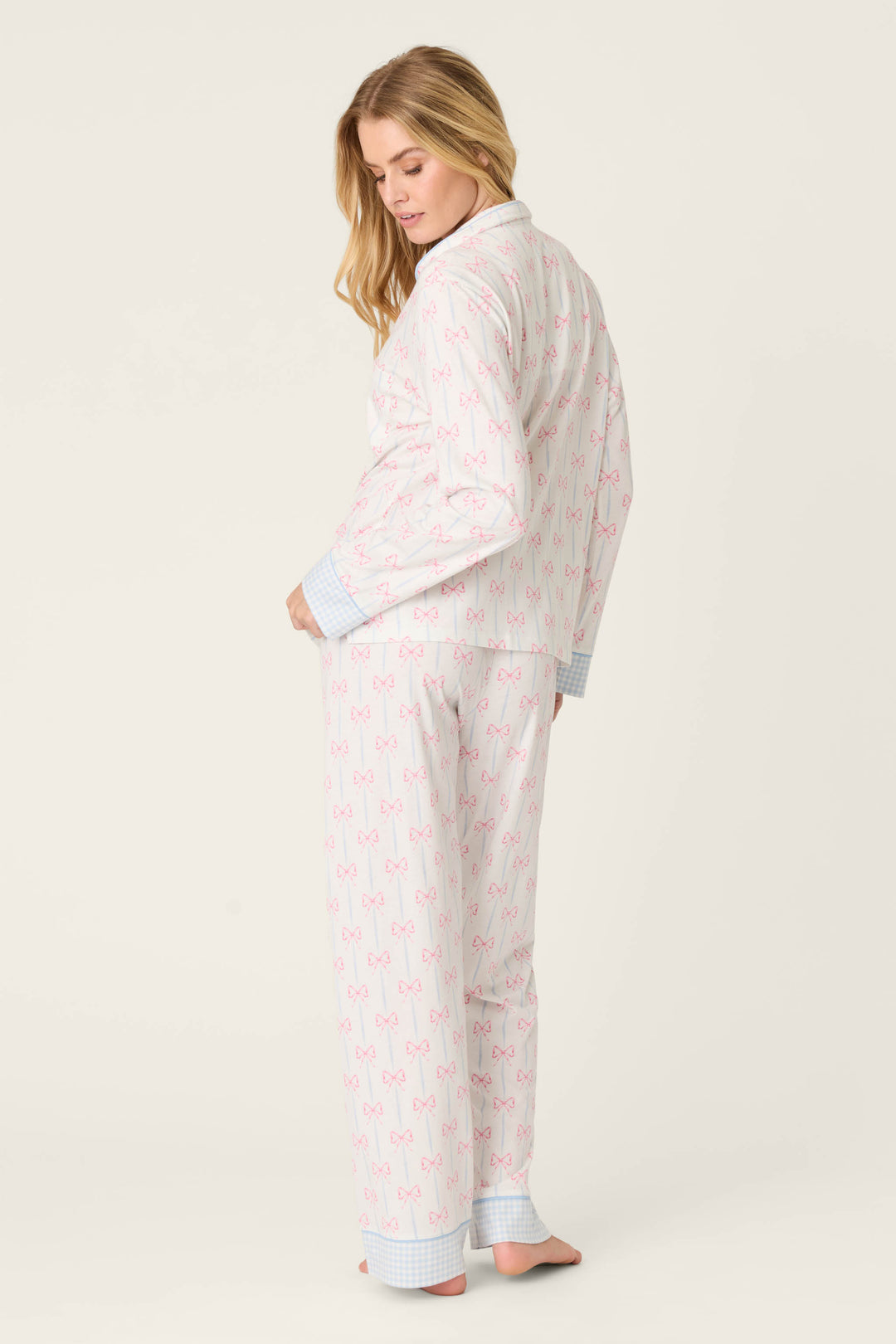 A barefoot person stands slightly turned, wearing the Playful Prints All Wrapped Up Long PJ Set by P.J. Salvage—white fabric with pink bows and blue plaid cuffs—against a plain off-white background.