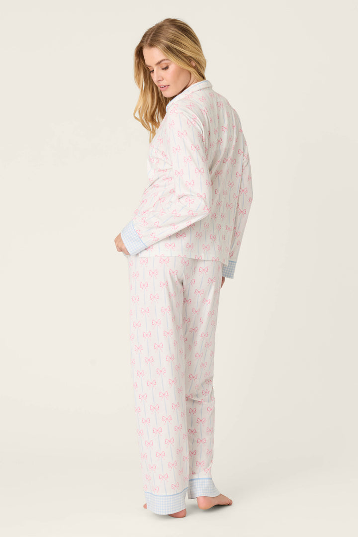 A barefoot person stands slightly turned, wearing the Playful Prints All Wrapped Up Long PJ Set by P.J. Salvage—white fabric with pink bows and blue plaid cuffs—against a plain off-white background.