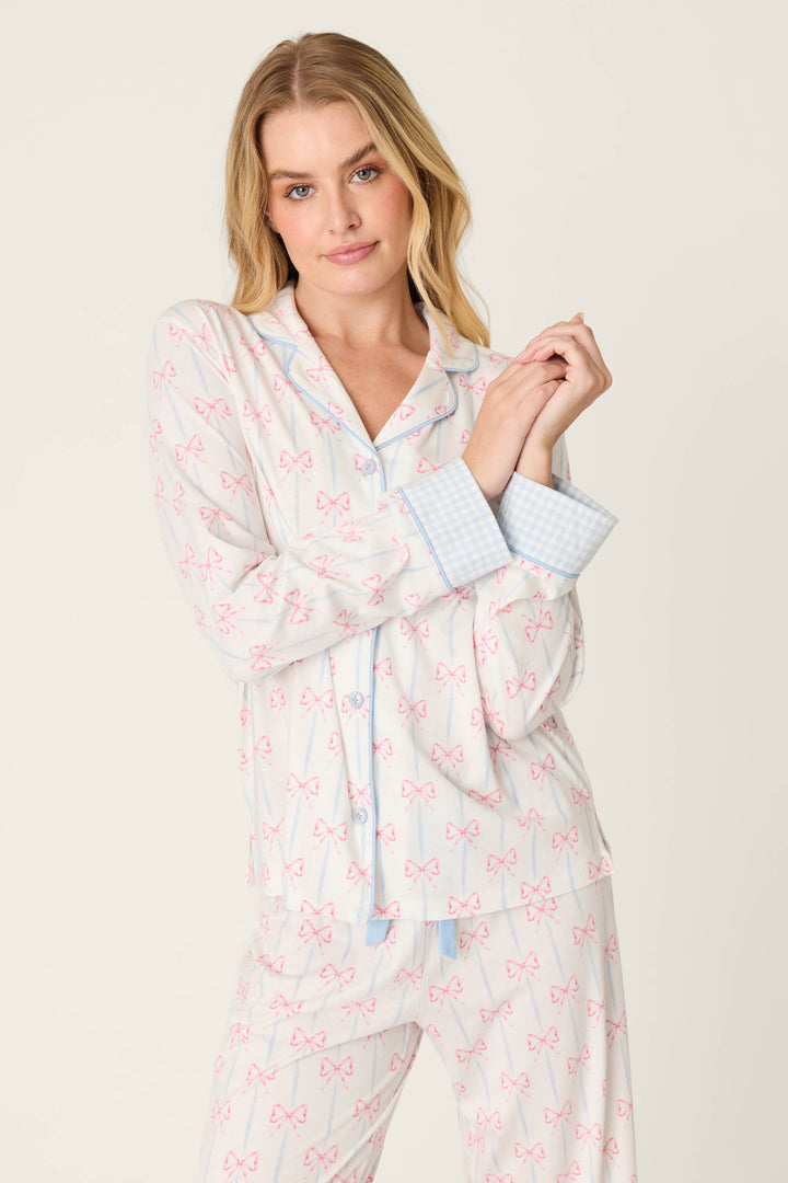 A model wears the Playful Prints All Wrapped Up Long PJ Set by P.J. Salvage, featuring a women’s bow-print design with a button-front shirt, pink bows, and light blue checkered cuffs, standing against a plain background and looking at the camera.