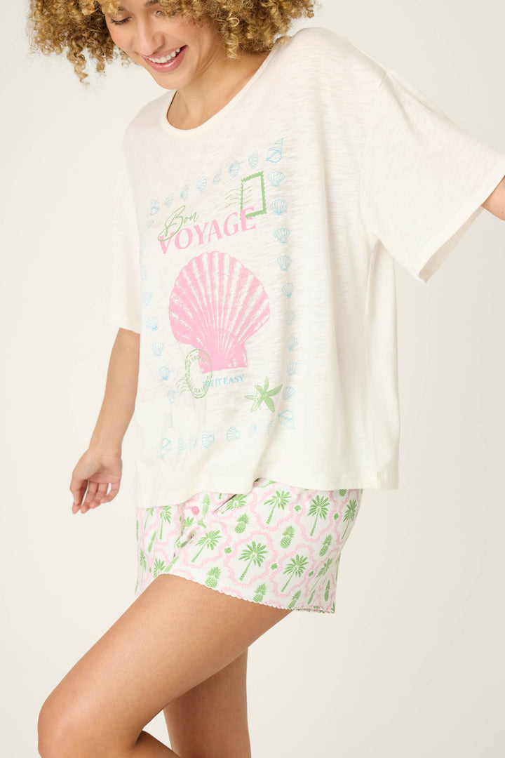 A person wears the P.J. Salvage Playful Prints Bon Voyage Graphic Tee—a loose white cotton-modal slub jersey top with a pink shell graphic—paired with pink and green patterned shorts, smiling and looking down.