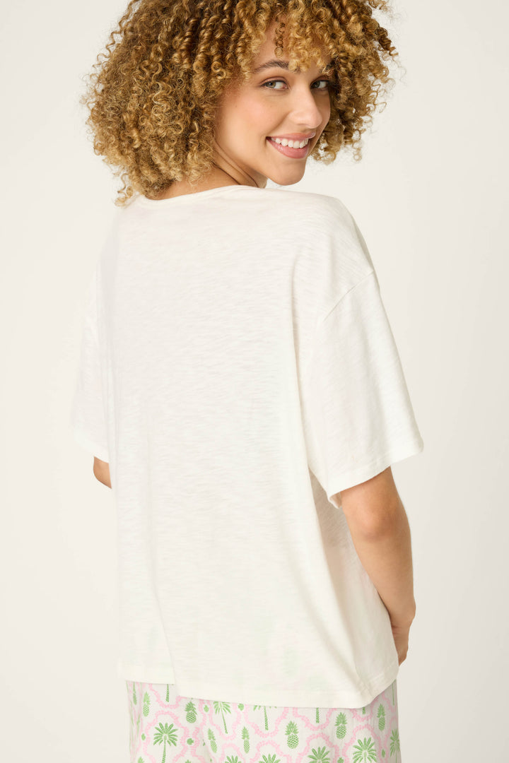 A person with curly hair smiles over their shoulder, wearing the P.J. Salvage Playful Prints Bon Voyage Graphic Tee and light pink shorts with a green and pink plant pattern, standing against a plain background.