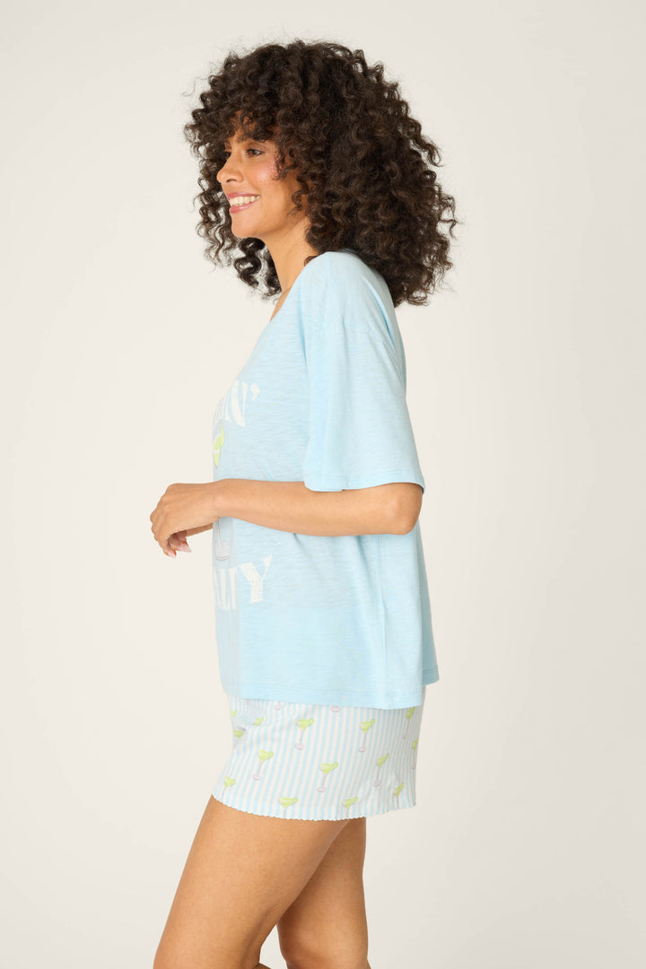 A woman with curly hair stands in profile, smiling, wearing the P.J. Salvage Playful Prints Feeling Salty Graphic Tee and matching striped shorts, exuding vacation vibes against a plain light background.