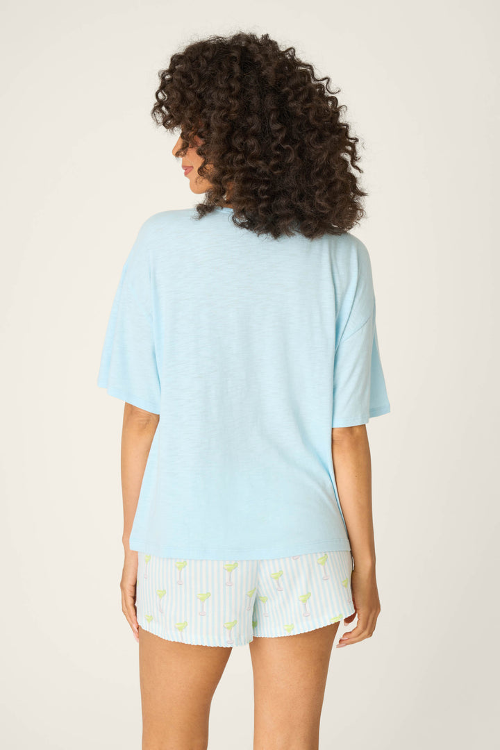 A person with curly hair stands with their back to the camera, wearing the P.J. Salvage Playful Prints Feeling Salty Graphic Tee and patterned white shorts, channeling vacation vibes against a plain background.