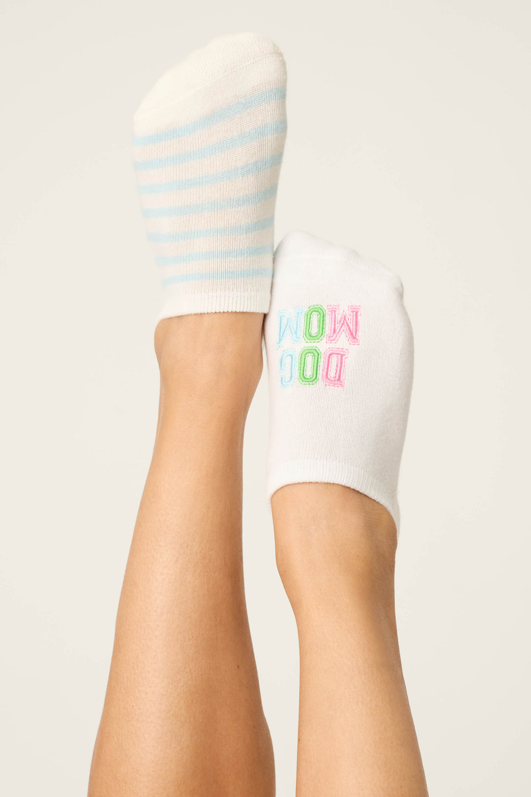 Feet raised in the air sport mismatched P.J. Salvage Pupcation Palms Dog Mom 2-Pack Cushion Liner Socks: one with white and light blue stripes, the other with a colorful mirrored "WOW DOG"—ideal for playful dog moms.