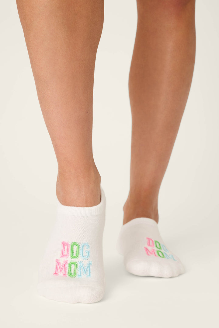A person stands on a light surface wearing P.J. Salvage’s Pupcation Palms Dog Mom 2-Pack Cushion Liner Socks, featuring “DOG MOM” embroidered in colorful letters. Only their lower legs and feet are visible, highlighting the playful design.