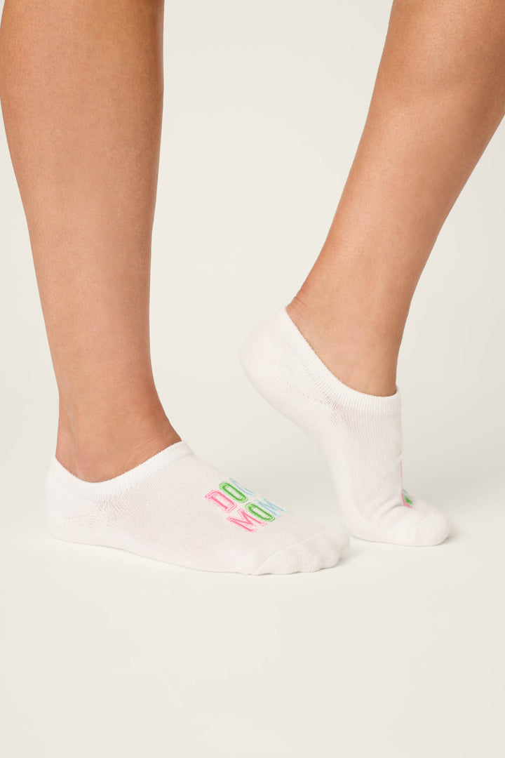 A person wears P.J. Salvage Pupcation Palms Dog Mom 2-Pack Cushion Liner Socks—white, low-profile socks with colorful embroidered text—while standing on a light surface, with only their lower legs and feet visible.