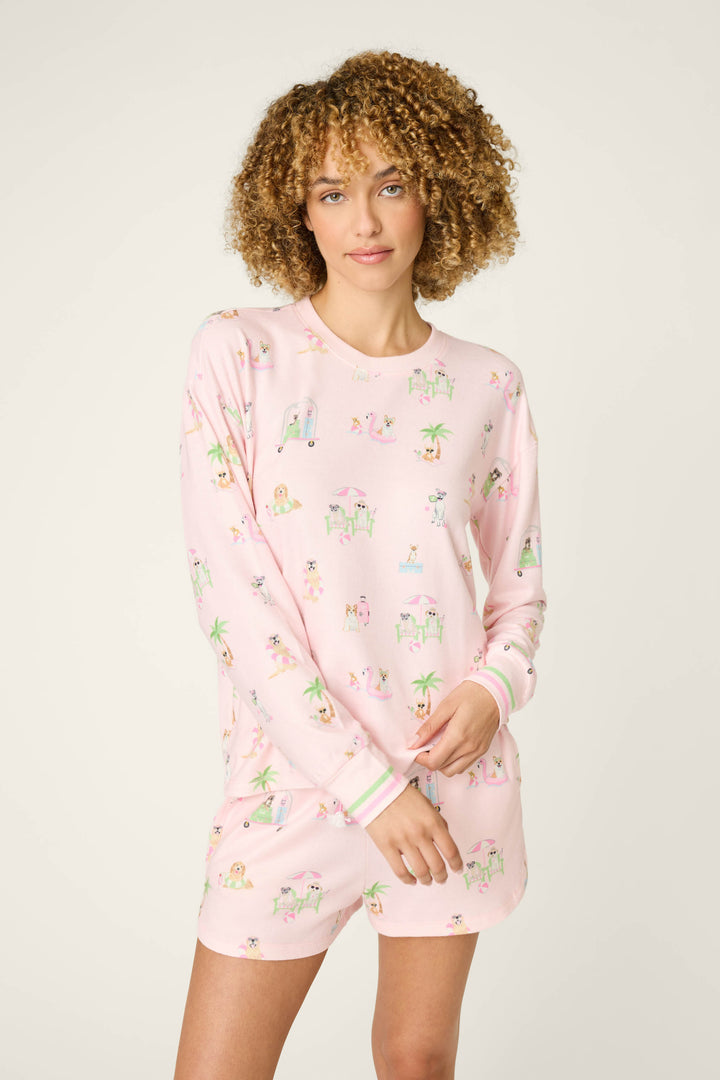 A woman with curly hair models the Pupcation Palms Sunnies & Sunshine Long Sleeve Top by P.J. Salvage, a light pink pajama featuring vacation dog and palm tree prints, exuding cozy comfort against a plain background.