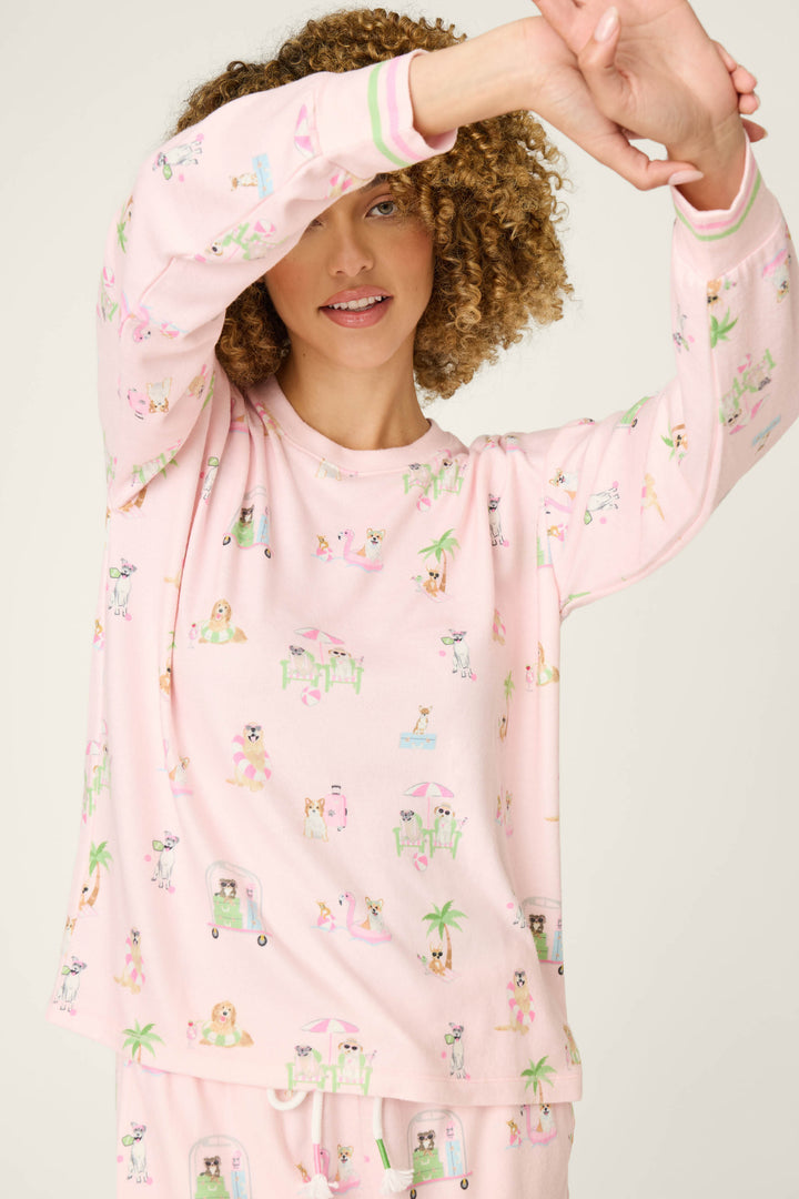 A person with curly hair in a P.J. Salvage Pupcation Palms Sunnies & Sunshine Long Sleeve Top, featuring light pink fabric with colorful prints, stands against a plain background, raising one arm to partially cover their face.