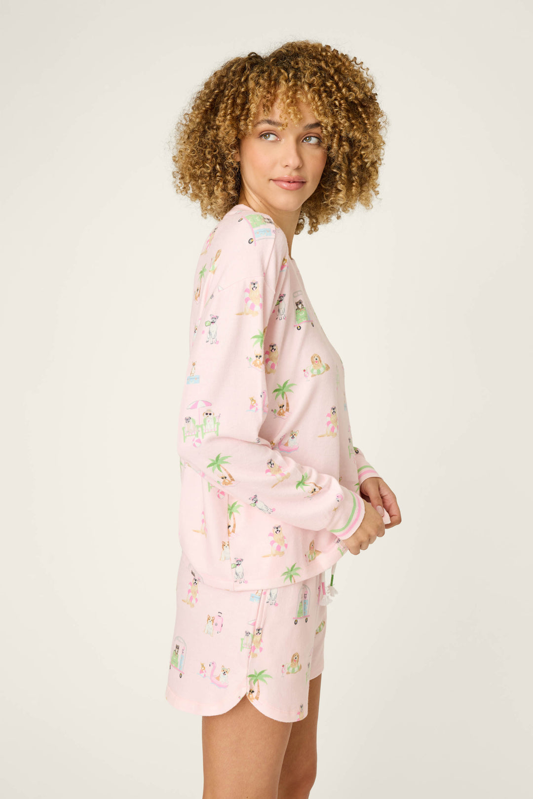 A person with curly hair stands sideways in a cozy P.J. Salvage Pupcation Palms Sunnies & Sunshine Long Sleeve Top, featuring colorful cartoon prints, against a plain light background.