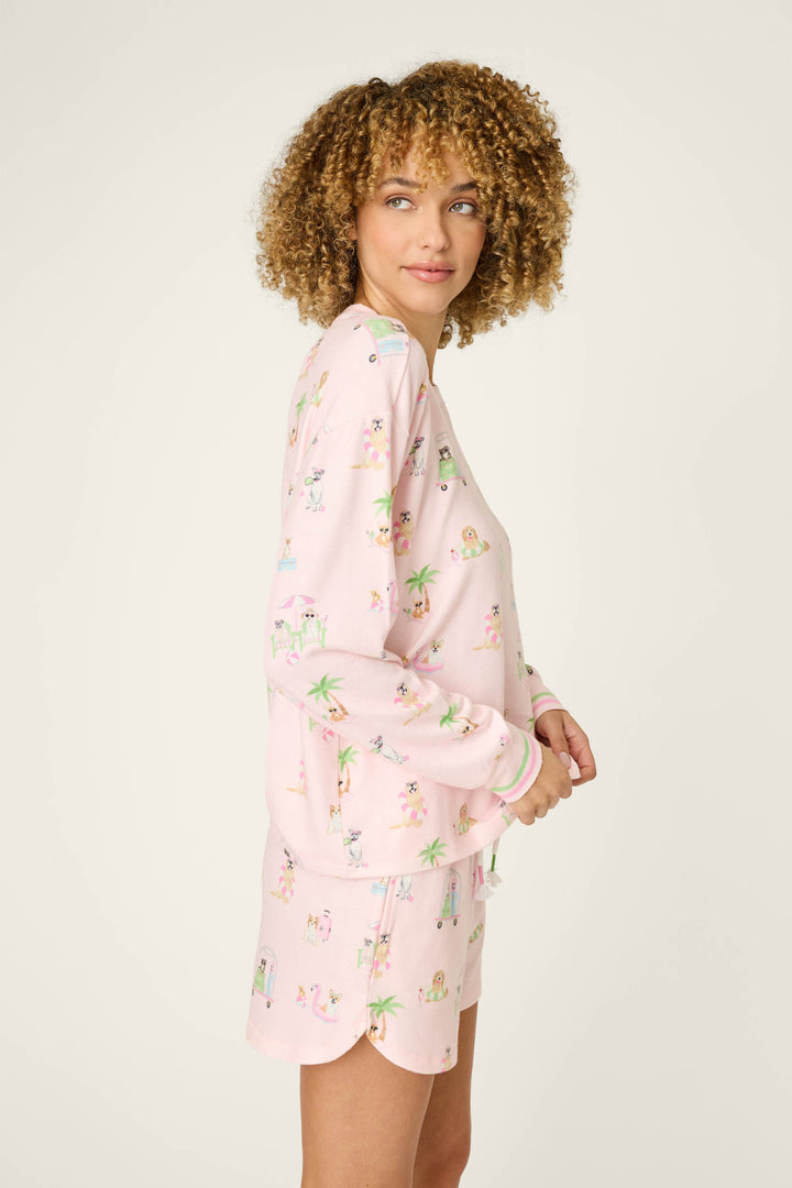 A person with curly hair stands sideways in a cozy P.J. Salvage Pupcation Palms Sunnies & Sunshine Long Sleeve Top, featuring colorful cartoon prints, against a plain light background.