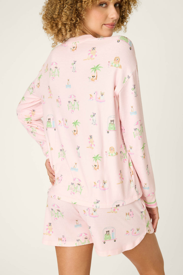 A person with curly hair wears the P.J. Salvage Pupcation Palms Sunnies & Sunshine Long Sleeve Top in light pink, featuring a playful vacation dog print. Turned to the side, they look over their shoulder, radiating cozy comfort.