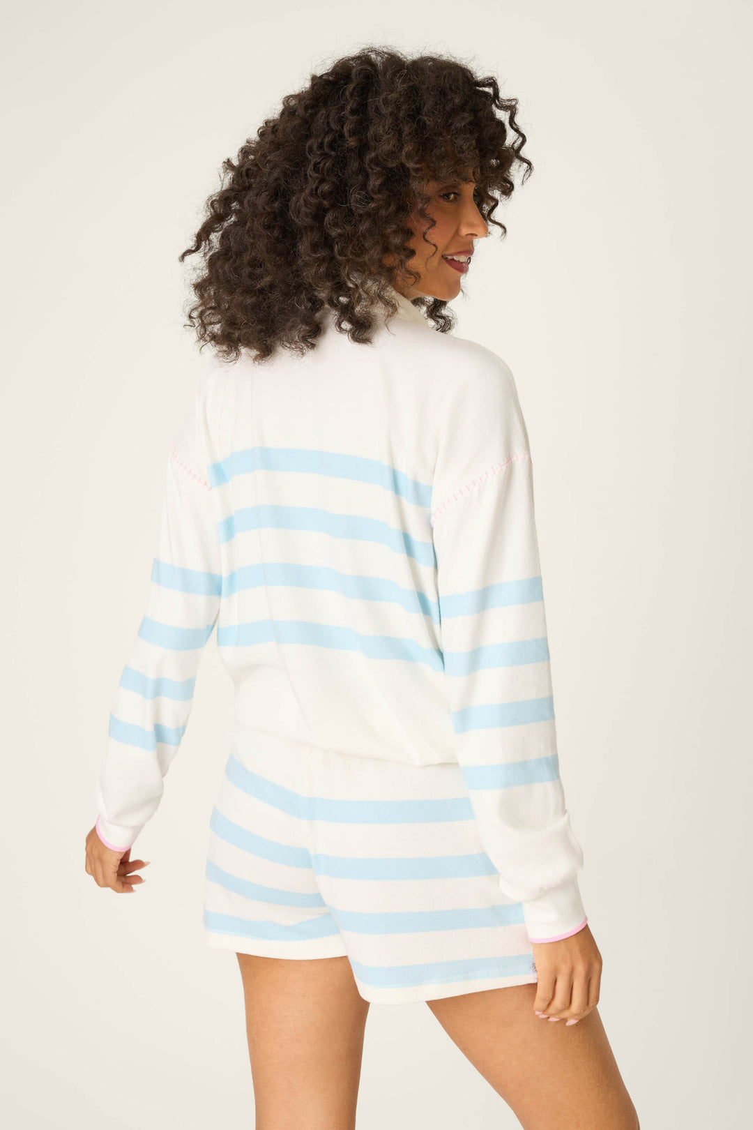 A person with curly hair is shown from the back, wearing the P.J. Salvage Pupcation Palms Ocean Stripe Quarter Zip Pullover Sweater, featuring a relaxed silhouette and light blue stripes, against a plain light background.