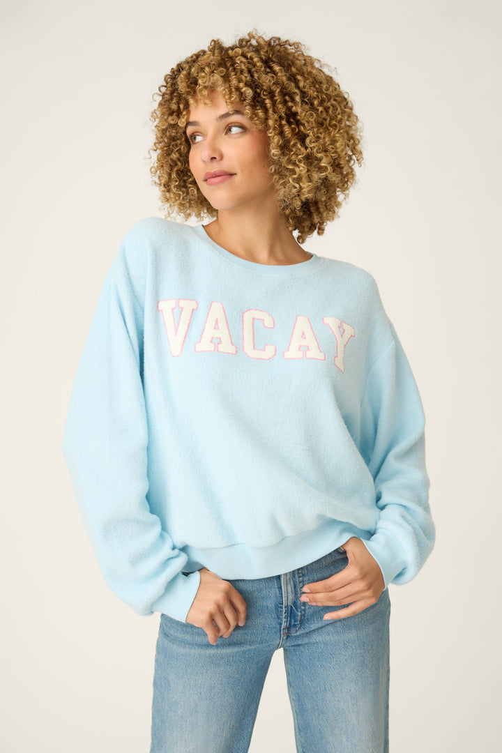 A person wearing the P.J. Salvage Pupcation Palms Vacay Pullover Sweater with bold Vacay lettering and blue jeans, standing against a plain light background.