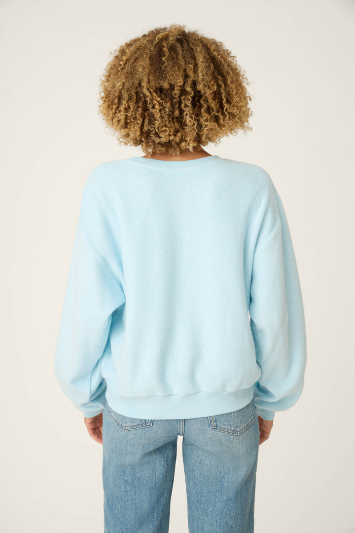 A person with curly hair is shown from the back, wearing the P.J. Salvage Pupcation Palms Vacay Pullover Sweater and light blue jeans, standing against a plain, light-colored background.