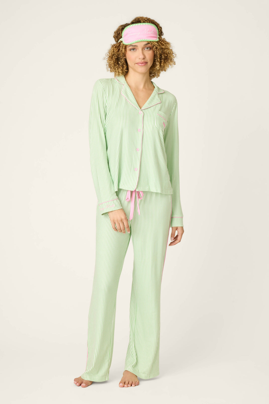 A woman stands barefoot facing forward in the P.J. Salvage Pupcation Palms Kiwi Stripe 3-Piece PJ Set, featuring green and white stripes with pink accents and a matching sleep mask, against a plain light background.