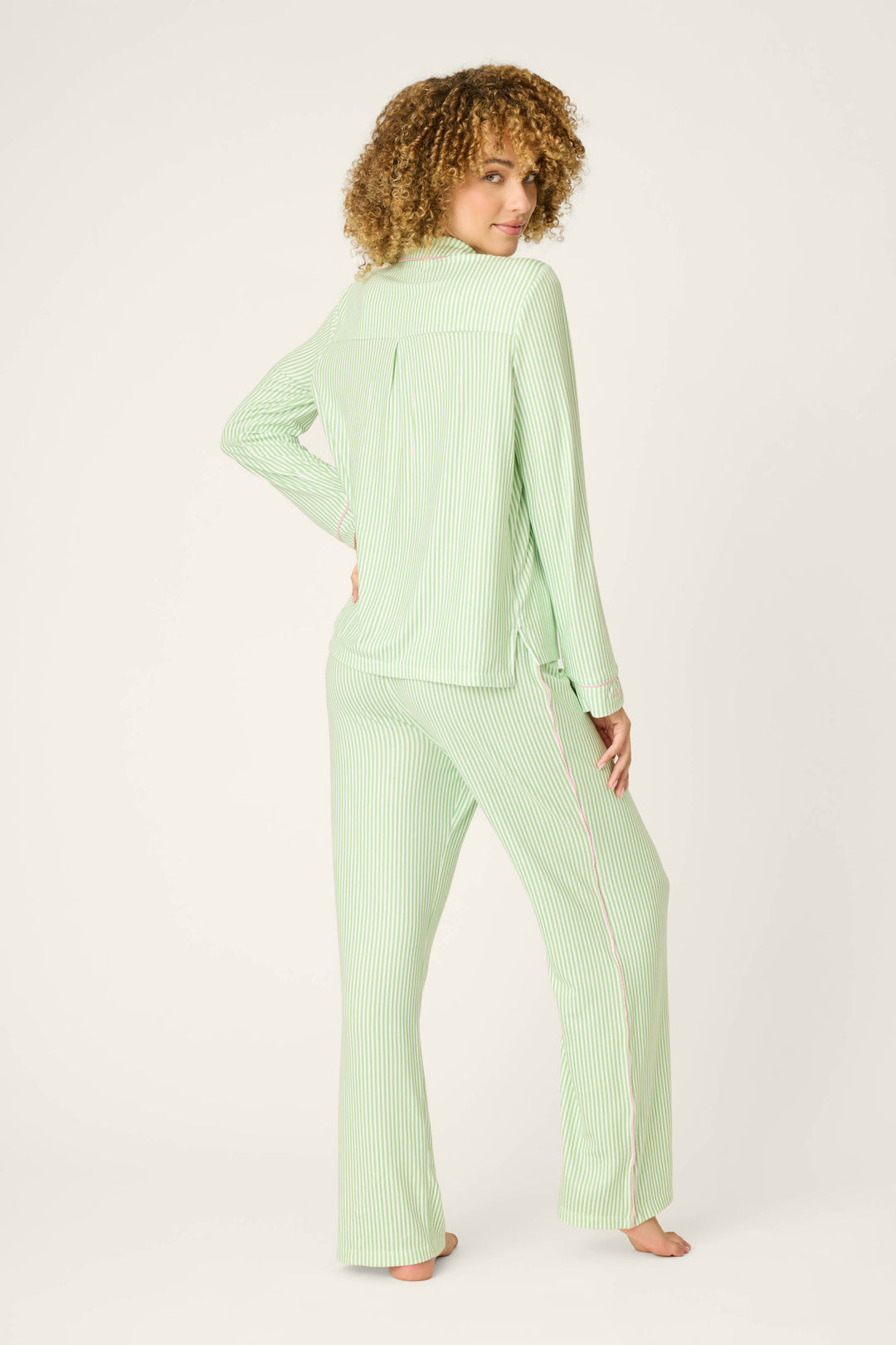 A person stands barefoot, glancing over their shoulder while wearing the P.J. Salvage Pupcation Palms Kiwi Stripe 3-Piece PJ Set, featuring green stripes and long sleeves, against a plain background.