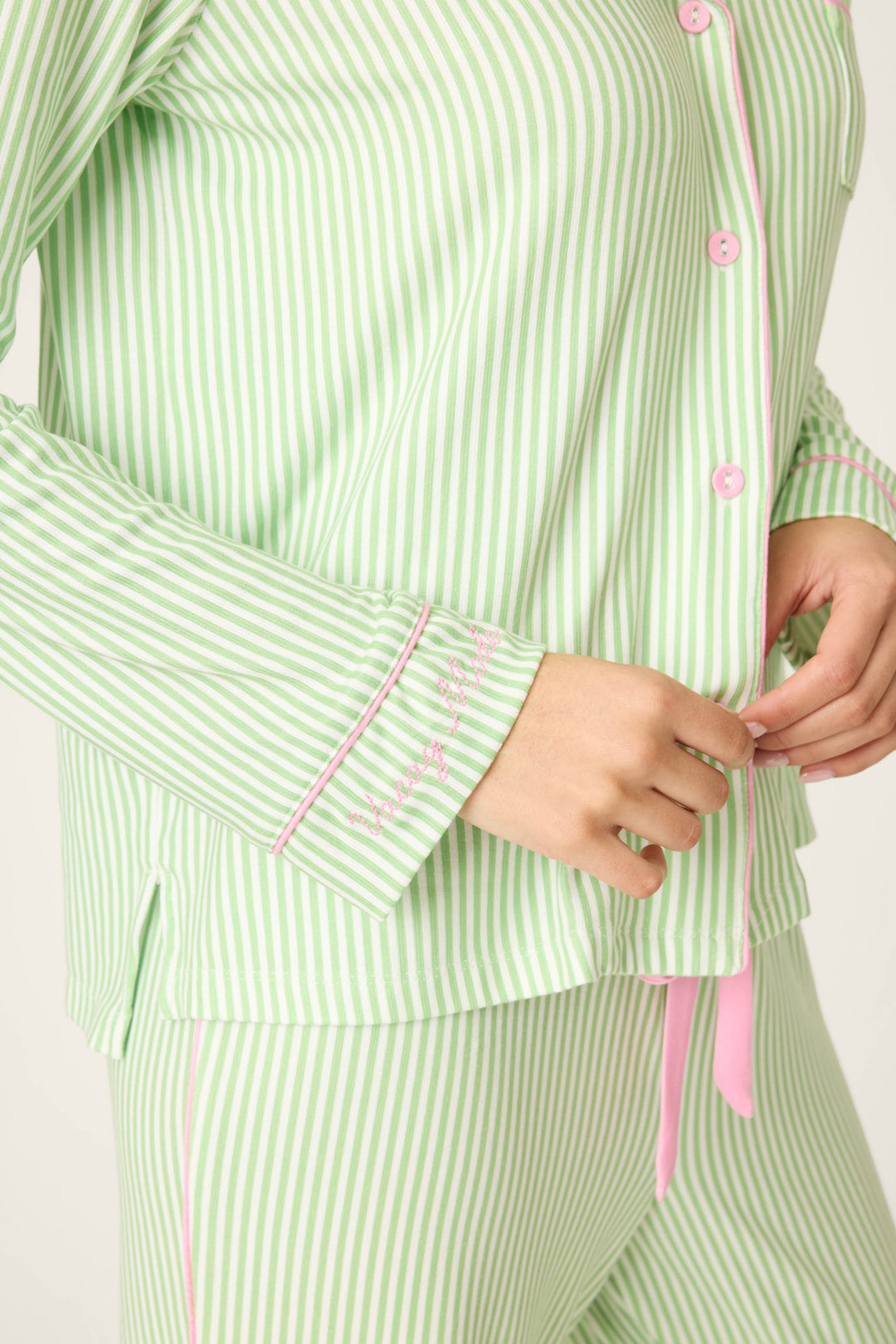A person wears the P.J. Salvage Pupcation Palms Kiwi Stripe 3-Piece PJ Set, shown from shoulders to hips. One hand adjusts the pajama top, highlighting pink trim, buttons, and delicate embroidery on the cuff.