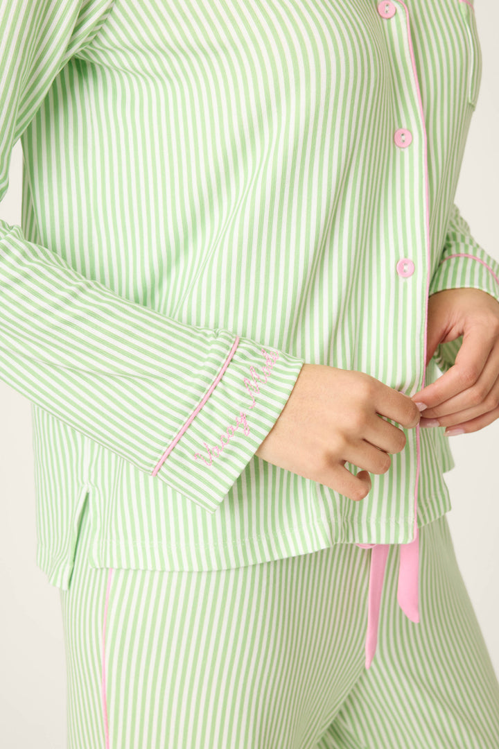 A person wears the P.J. Salvage Pupcation Palms Kiwi Stripe 3-Piece PJ Set, shown from shoulders to hips. One hand adjusts the pajama top, highlighting pink trim, buttons, and delicate embroidery on the cuff.