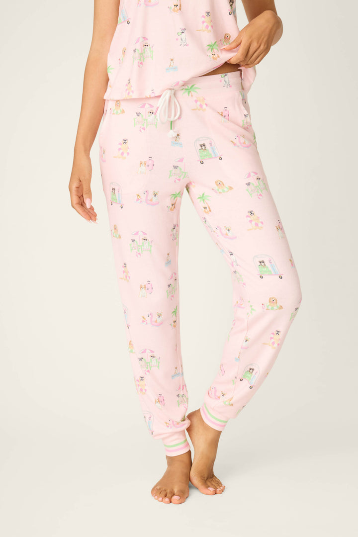 The lower half of a person is shown barefoot against a plain background, wearing P.J. Salvage Pupcation Palms Sunnies & Sunshine Banded Pants—light pink joggers with a white drawstring waist and colorful prints.