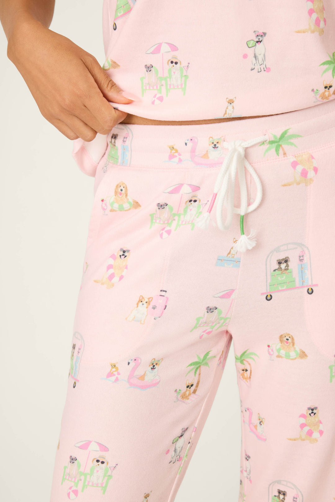 A person wears P.J. Salvage Pupcation Palms Sunnies & Sunshine Banded Pants and a matching top in soft peach jersey with playful dog prints, palm trees, and pool floats. The comfy set features a drawstring waist; only torso and part of one arm show.