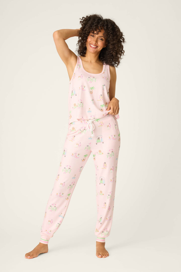 A curly-haired person smiles barefoot in P.J. Salvage’s Pupcation Palms Sunnies & Sunshine Banded Pants set—light pink jersey pajamas with a colorful print, sleeveless top, and jogger pants with a drawstring waist—against a plain light background.