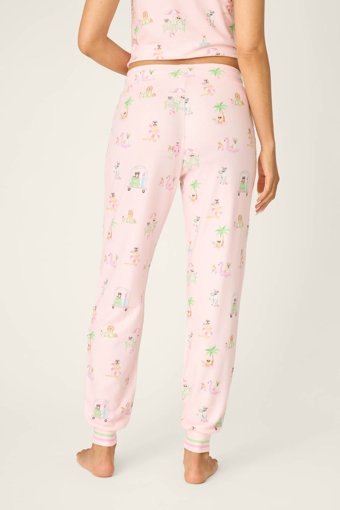 A person stands barefoot facing away, wearing P.J. Salvage Pupcation Palms Sunnies & Sunshine Banded Pants, featuring a drawstring waist and playful animal, palm tree, and vehicle prints on light pink fabric with a matching top.