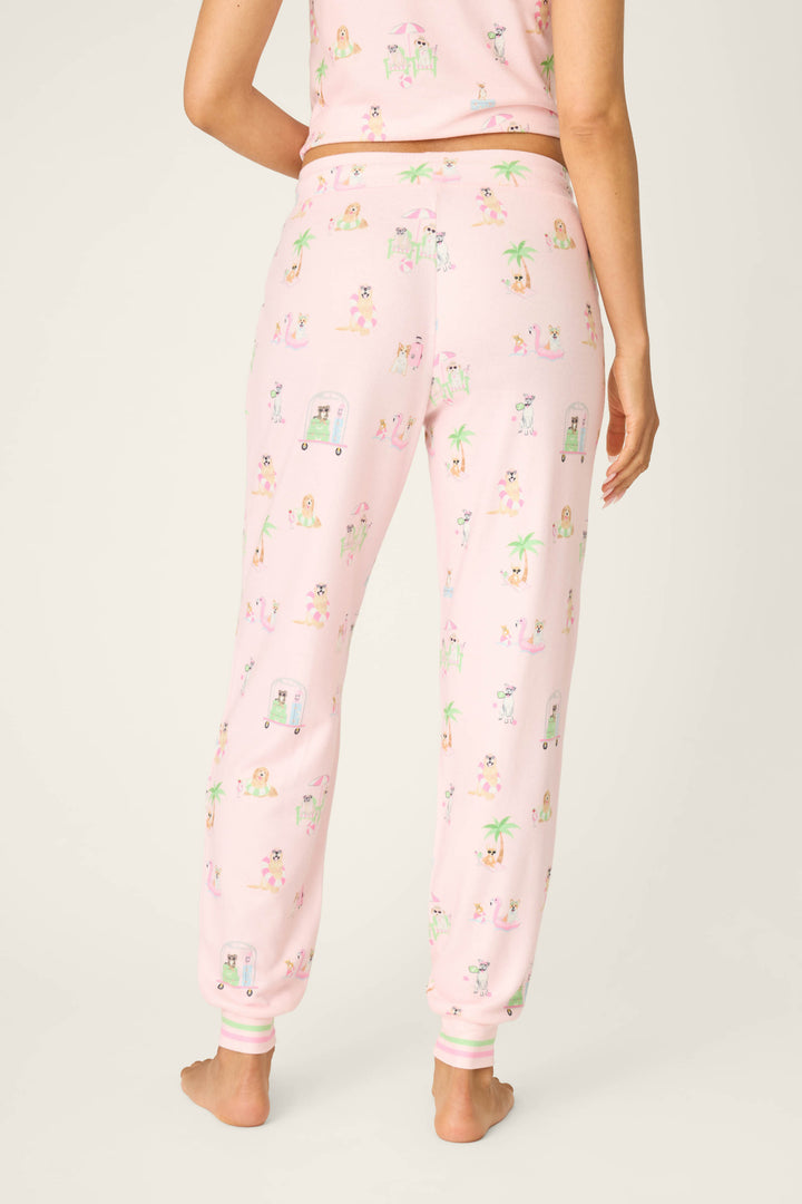 A person stands barefoot facing away, wearing P.J. Salvage Pupcation Palms Sunnies & Sunshine Banded Pants, featuring a drawstring waist and playful animal, palm tree, and vehicle prints on light pink fabric with a matching top.