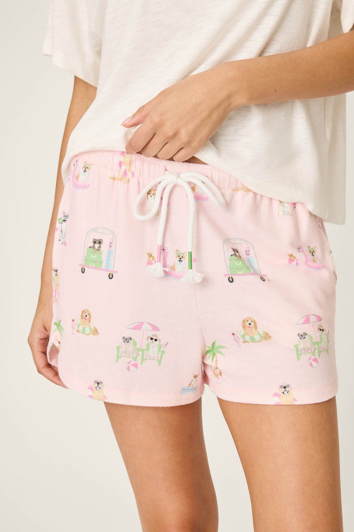A person wears P.J. Salvage Pupcation Palms Sunnies & Sunshine PJ Shorts in a runner-style fit, showcasing illustrated dogs, palm trees, and beaches on peachy jersey fabric; only their lower torso and upper legs are visible.