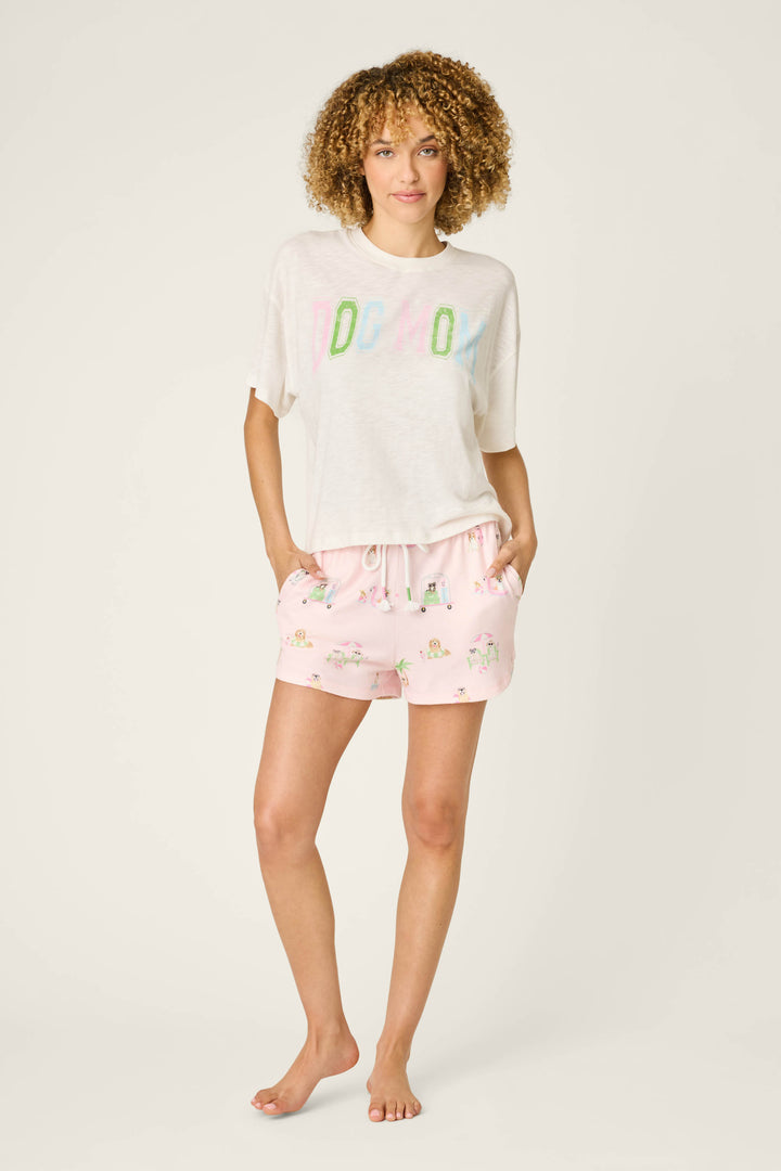 A woman with curly hair stands barefoot against a light background, wearing P.J. Salvage’s Pupcation Palms Sunnies & Sunshine PJ Shorts in peach and a loose white T-shirt with colorful text, hands in her pockets.