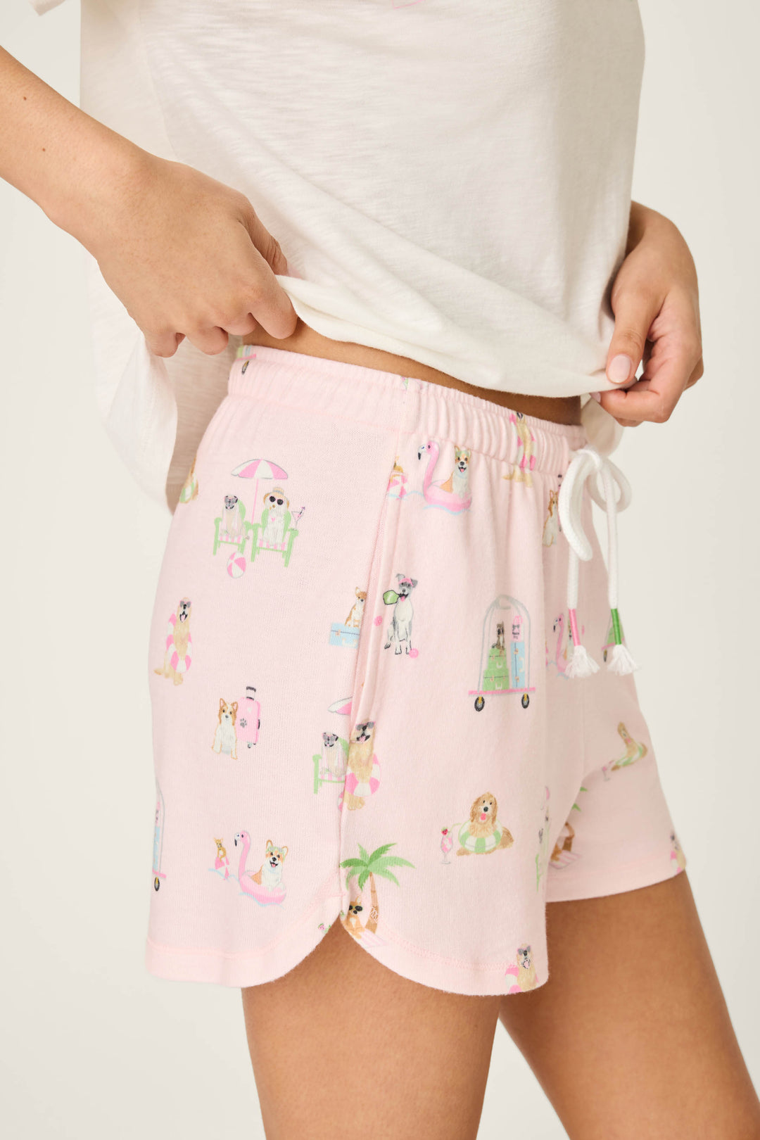 Wearing P.J. Salvage's Pupcation Palms Sunnies & Sunshine PJ Shorts—light pink with a playful dog print and white drawstring—a person stands in a white tee, hand on hip, showing off the runner-style vacation lounge look.