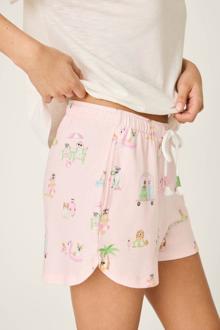 Wearing P.J. Salvage's Pupcation Palms Sunnies & Sunshine PJ Shorts—light pink with a playful dog print and white drawstring—a person stands in a white tee, hand on hip, showing off the runner-style vacation lounge look.