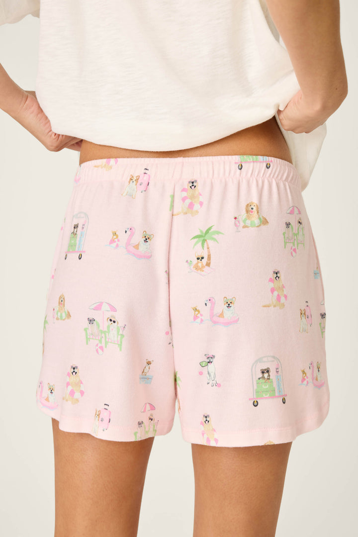 Wearing P.J. Salvage's Pupcation Palms Sunnies & Sunshine PJ Shorts, featuring playful pup prints with umbrellas, floaties, and palm trees in a runner-style fit, they stand with hands on hips.