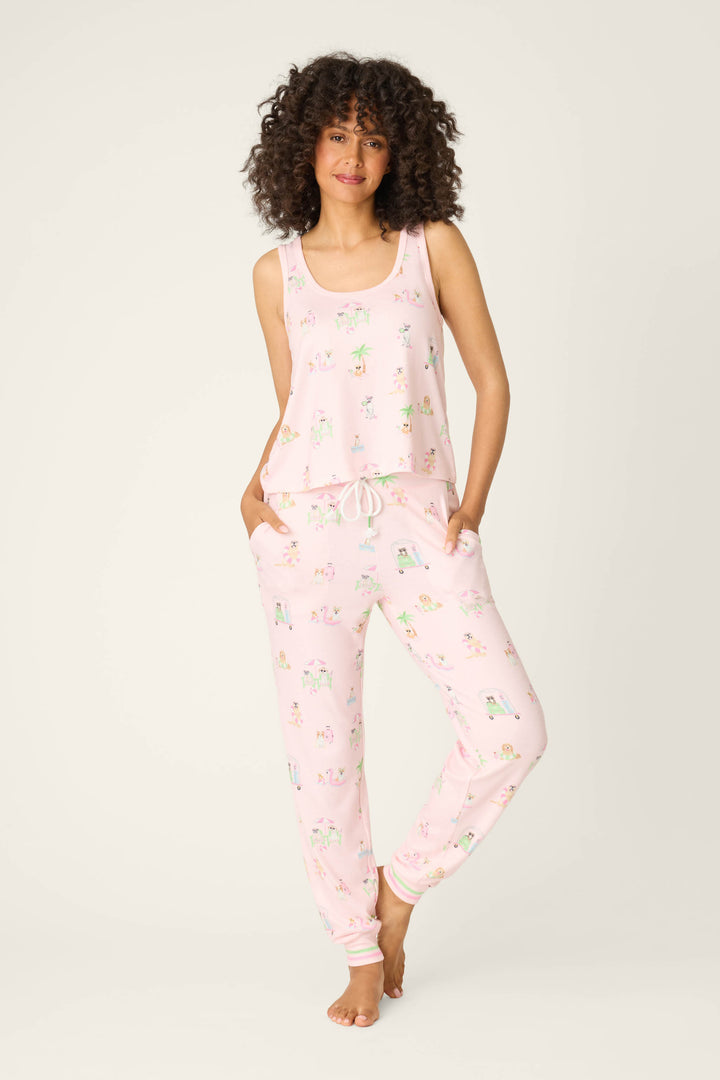 A person with curly hair stands barefoot, wearing the P.J. Salvage Pupcation Palms Sunnies & Sunshine Long PJ Set—a light pink tank top and drawstring pants with a playful print—hands in pockets against a plain light background.