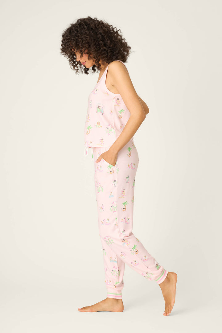 A woman with curly hair stands barefoot in profile, wearing the P.J. Salvage Pupcation Palms Sunnies & Sunshine Long PJ Set—a light pink tank and joggers with a playful print. The background is plain and light-colored.