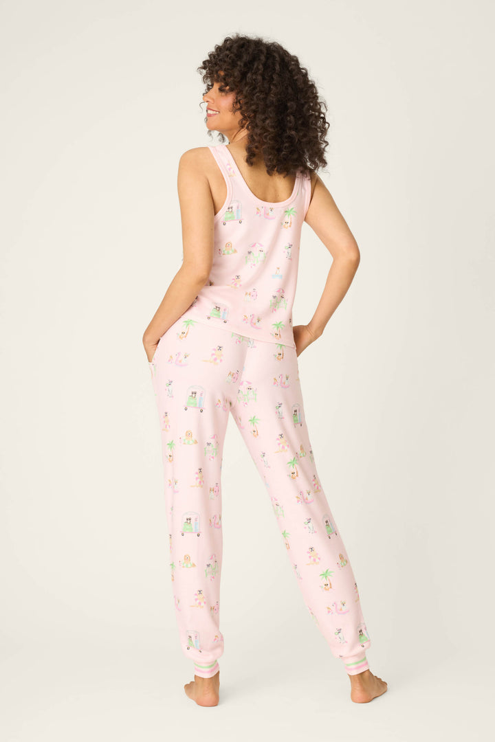 A person stands barefoot facing away, wearing the P.J. Salvage Pupcation Palms Sunnies & Sunshine Long PJ Set, a light pink lounge-ready pajama set with a tank and jogger pants featuring a colorful animal print. The background is off-white.