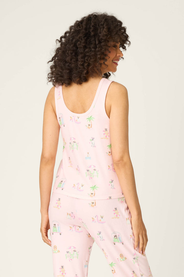 A woman with curly hair stands facing away, wearing the P.J. Salvage Pupcation Palms Sunnies & Sunshine Tank Top in light pink and matching printed pants featuring playful, colorful illustrations.