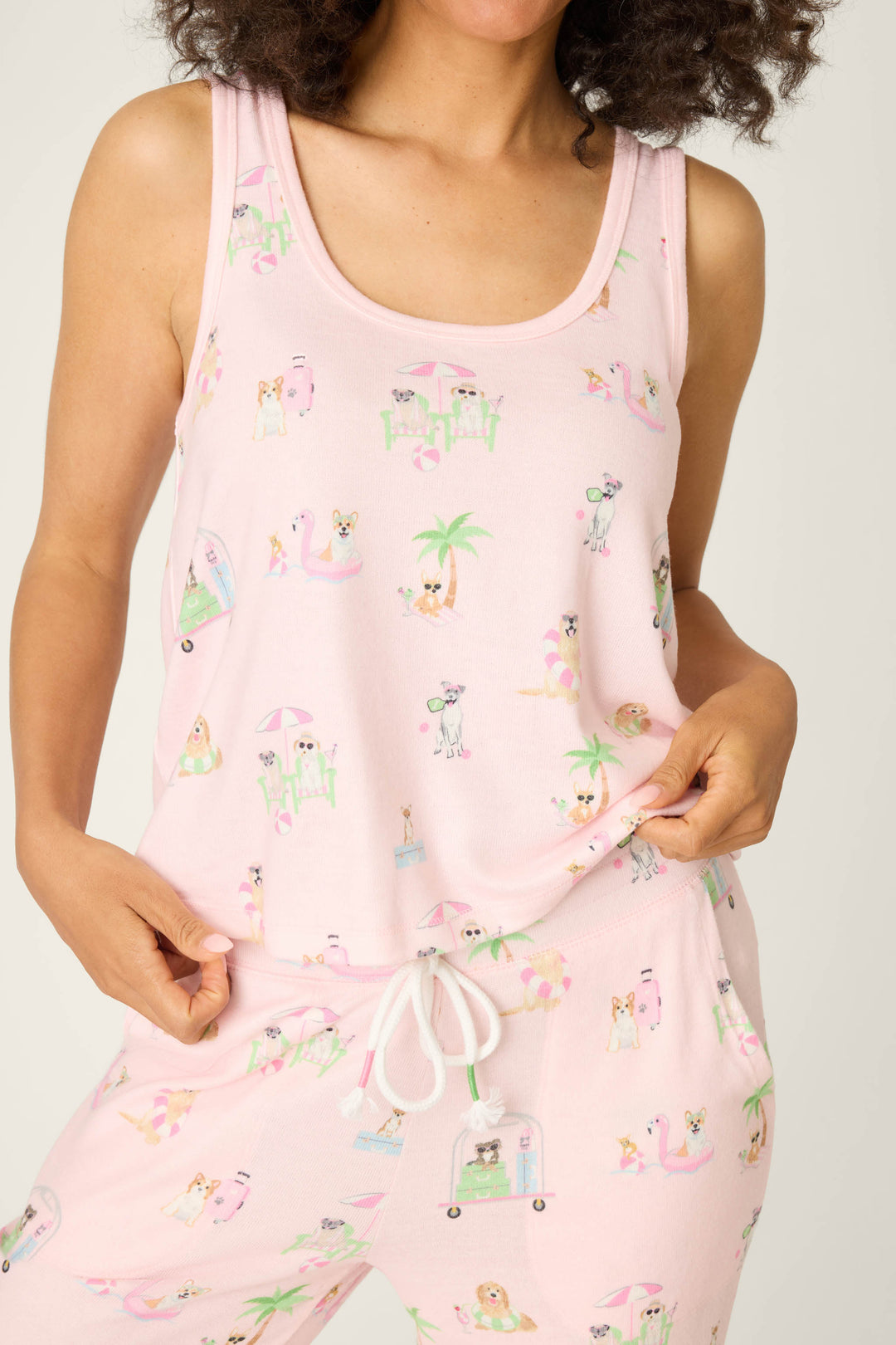 A person wears the P.J. Salvage Pupcation Palms Sunnies & Sunshine Tank Top—light pink, peachy jersey, sleeveless with a drawstring waist and playful prints of animals, palm trees, and beach scenes—while holding the hem.