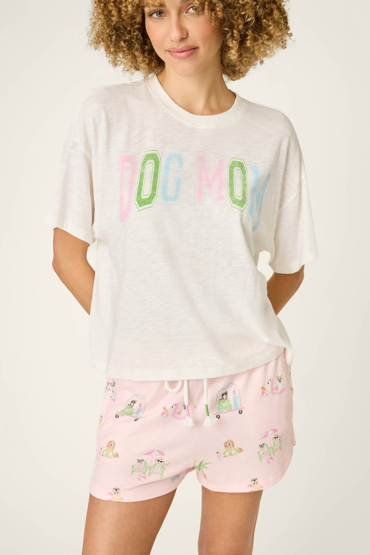 A woman with curly hair wears the P.J. Salvage Pupcation Palms Dog Mom Graphic Tee and light pink dog-and-beach print shorts, standing with her hands behind her back against a plain background.