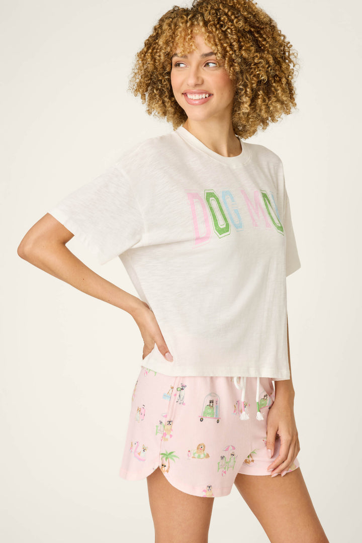 A curly-haired person wears the P.J. Salvage Pupcation Palms Dog Mom Graphic Tee with colorful text, paired with light pink pajama shorts featuring small prints, standing and smiling against a plain light background.