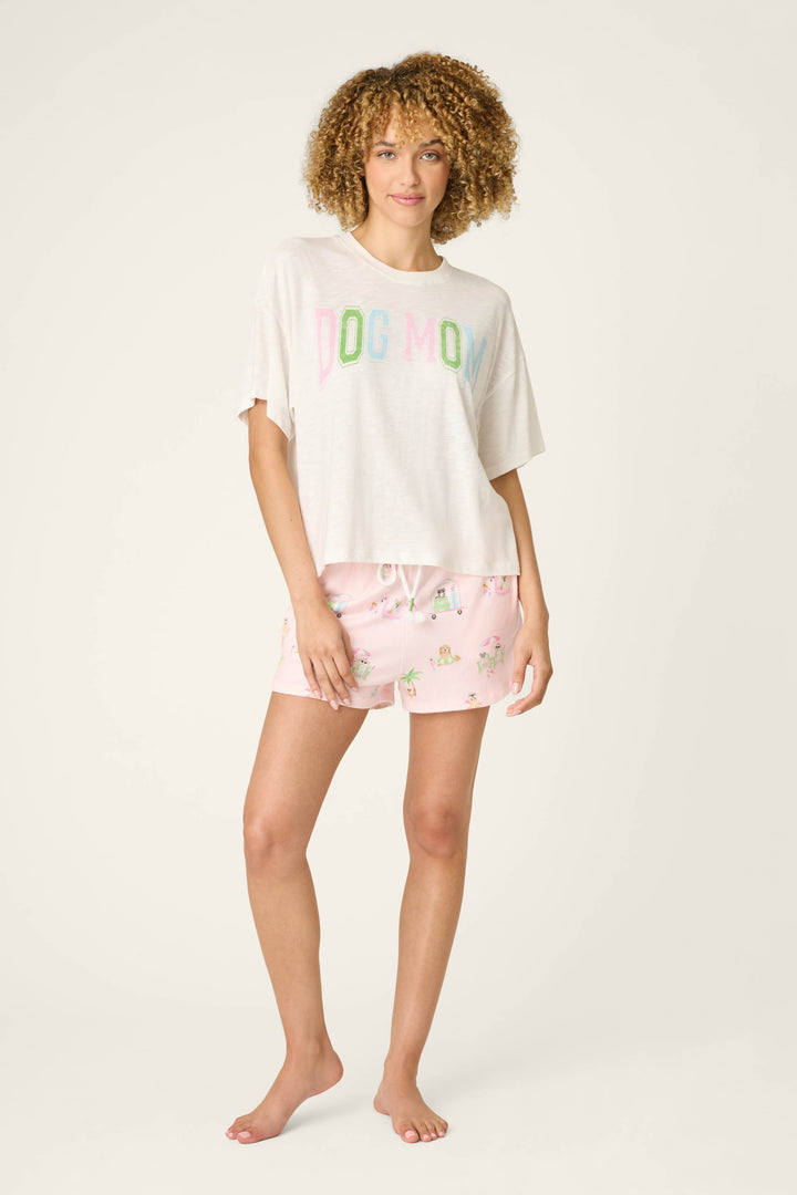 A woman with curly hair stands barefoot against a plain background, wearing the P.J. Salvage Pupcation Palms Dog Mom Graphic Tee with a relaxed crewneck and pink patterned drawstring shorts.