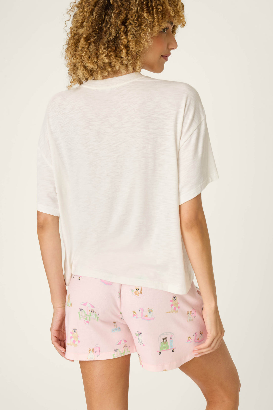 A person with curly hair is shown from behind wearing the P.J. Salvage Pupcation Palms Dog Mom Graphic Tee and light pink shorts with colorful prints, standing against a plain off-white background.