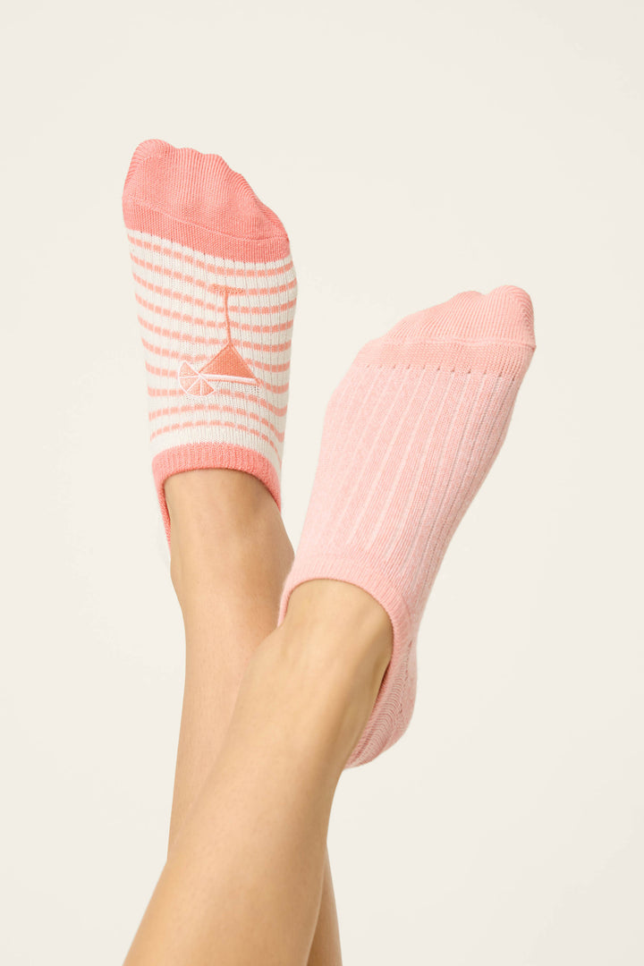 Two feet wear the P.J. Salvage Paloma Mama Sweet Sips 2-Pack No Show Sock Liners. The left foot sports a pink sock with white stripes and a graphic, while the right wears a solid light pink ribbed sock against a neutral background.
