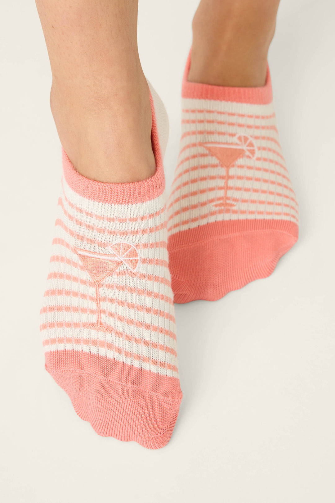 A pair of feet wear P.J. Salvage Paloma Mama Sweet Sips 2-Pack No Show Sock Liners, featuring pink and white stripes with a cocktail glass and citrus slice design on top, set against a plain light background.