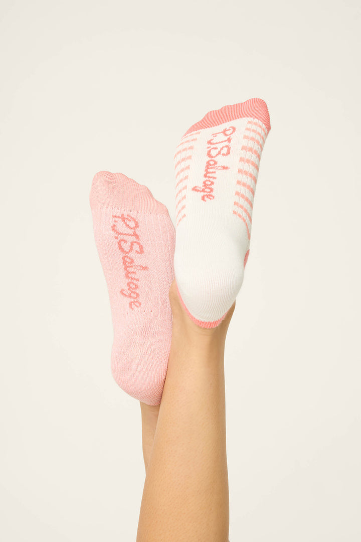 Two feet are raised against a plain background in mismatched Paloma Mama Sweet Sips 2-Pack No Show Sock Liners by P.J. Salvage—one solid pink, the other white with pink stripes—both featuring P.J. Salvage in pink text on the soles.