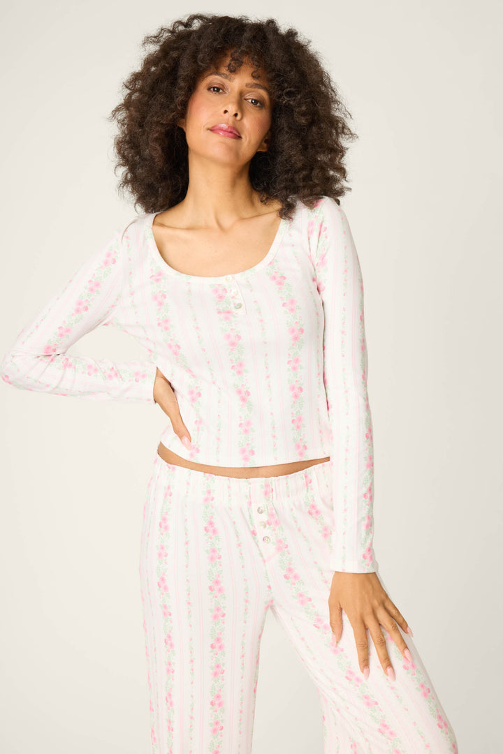A woman with curly hair wears the P.J. Salvage Roses Are Pink Petal Dream Pointelle Long Sleeve Top and matching pants with a light pink floral pattern, standing against a plain background with one hand on her hip.