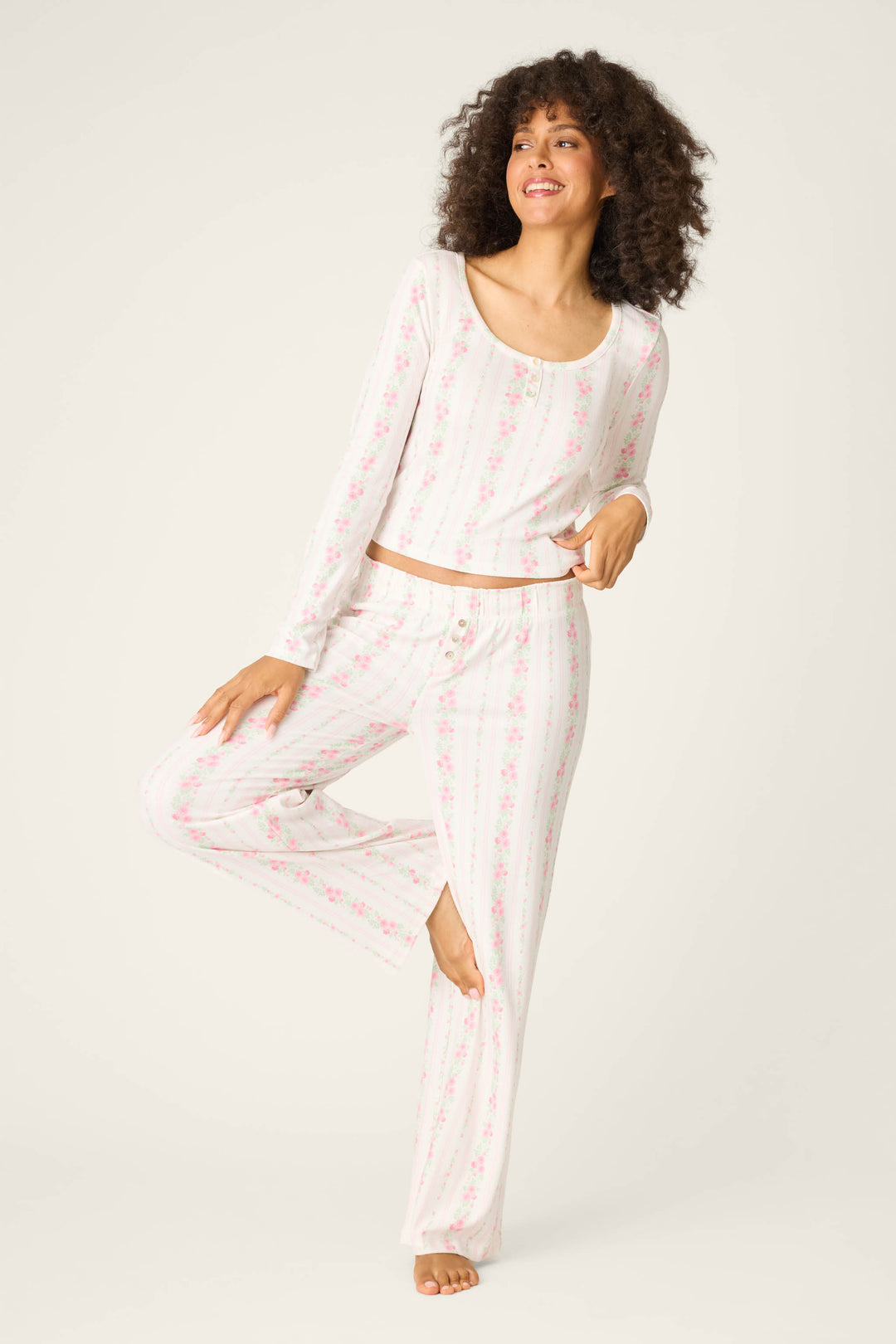 A person with curly hair stands barefoot on one leg, wearing the P.J. Salvage Roses Are Pink Petal Dream Pointelle Long Sleeve Top with a white and pink floral striped pattern, against a plain light background.
