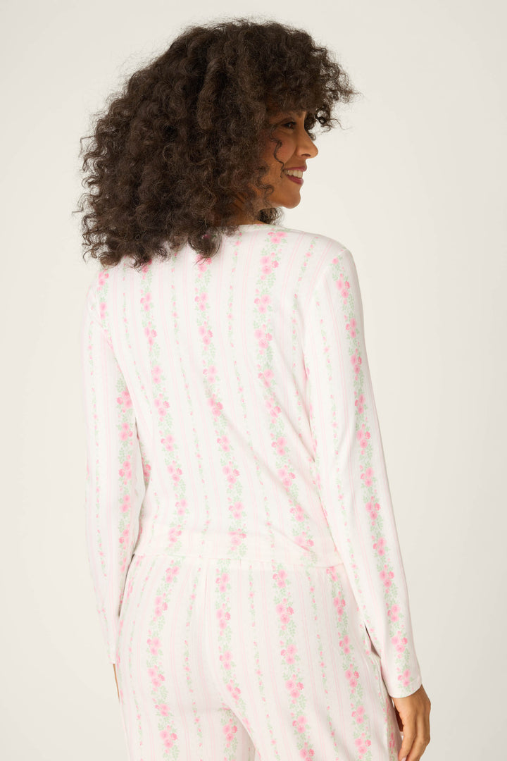 A person with curly hair is seen from the back, wearing the P.J. Salvage Roses Are Pink Petal Dream Pointelle Long Sleeve Top. The pajama top features a white base with a pink floral design against a plain, light background.