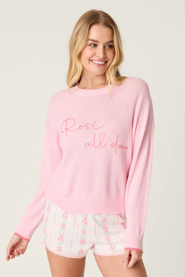 A smiling woman with long blonde hair wears the P.J. Salvage Roses Are Pink Rosé All Day Snuggle Sweater and white floral shorts, standing against a plain light background.