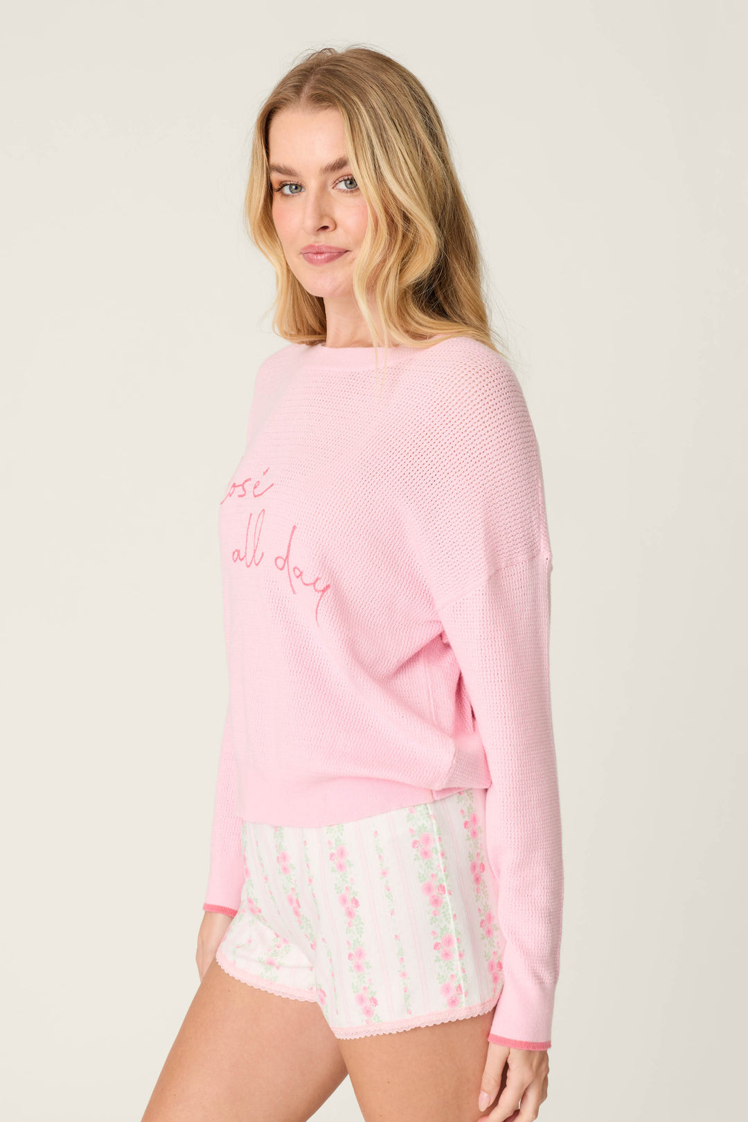 A woman with long blonde hair wears the P.J. Salvage "Roses Are Pink Rosé All Day Snuggle Sweater" in light pink, paired with white floral shorts, standing and facing the camera against a plain light background.